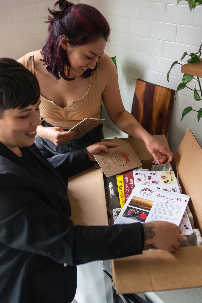 Two wine club members open a recent shipment of wine from Brooks Wine as well as educational tasting resources as gifts.
