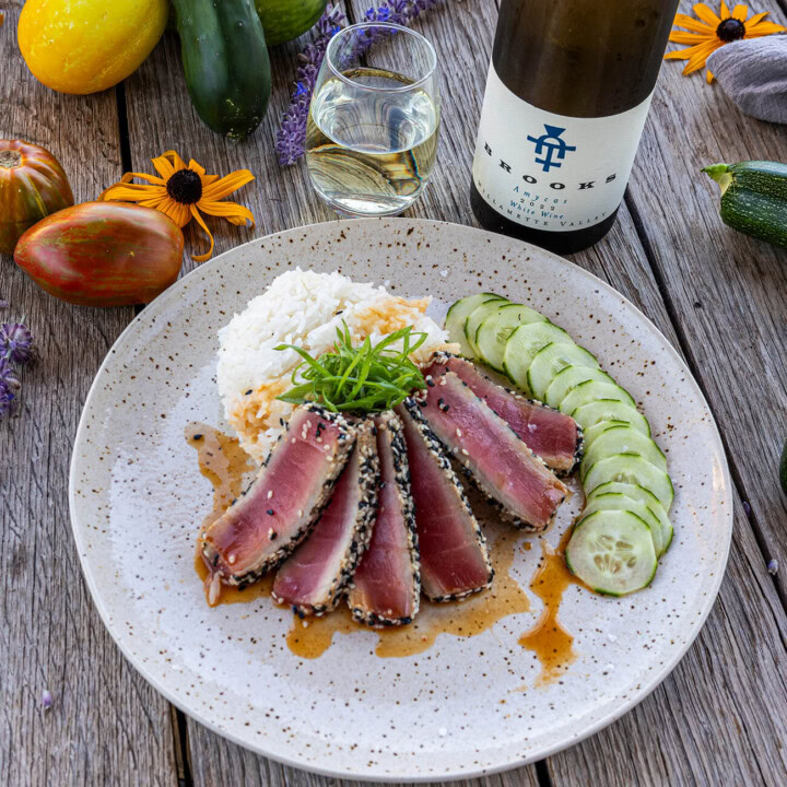A plate of seared ahi tuna crusted in sesame seeds alongside crisp cucumber dressed in a mirin and soy sauce glaze while paired with a refreshing glass of an Alsatian-inspired white wine blend.