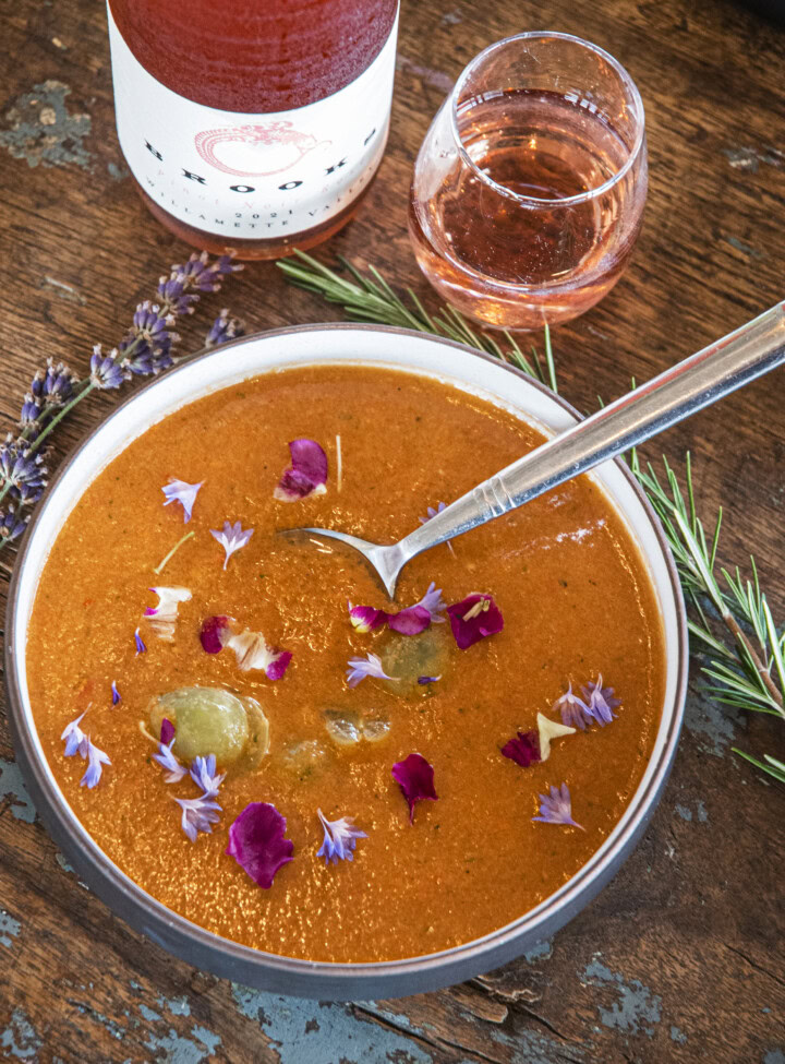 A nourishing soup made from vine ripened tomatoes is served with purple and maroon edible blossoms and a delicate rose wine.