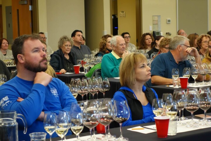A wine seminar is well attended with wine enthusiast students of all ages who are listening attentively to the speaker and sitting in rows of tables covered in black table clothes and set with white tasting mats, four glasses of white wine, and six glasses of red wine.