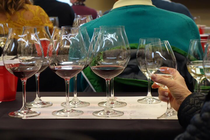 A student in a wine class grasps one of eight wine glasses resting on a white tasting mat to better examine the hue of the red wine she is tasting.