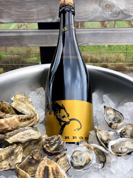 A bottle of sparkling wine is prominently displayed inside a silver ice bucket filled with chipped ice and multiple shucked oysters as an appetizer to dinner.