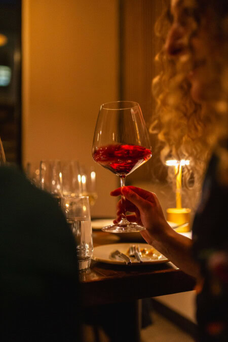 A dinner party guest swirls a glass of Pinot Noir in an elegant stemmed glass. Her face's side profile is partially hidden by her long, curly, blonde hair. The dinner table is in the background with dim lights set on the table.