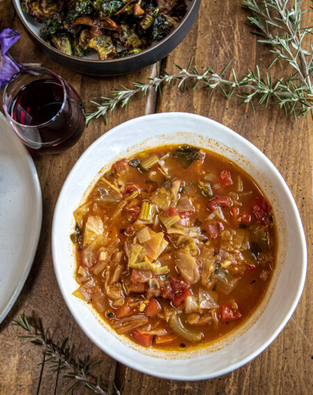 A close up photo of a wooden table set with rosemary sprigs and a white bowl of hearty tomato and cabbage stew is prominently displayed next to a glass of Pinot Noir.