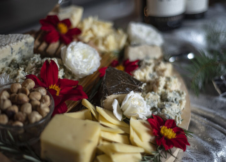 A wine and cheese pairing boasts a round wooden board with a large selection of unique, artisan cheeses and festive red and white flowers.