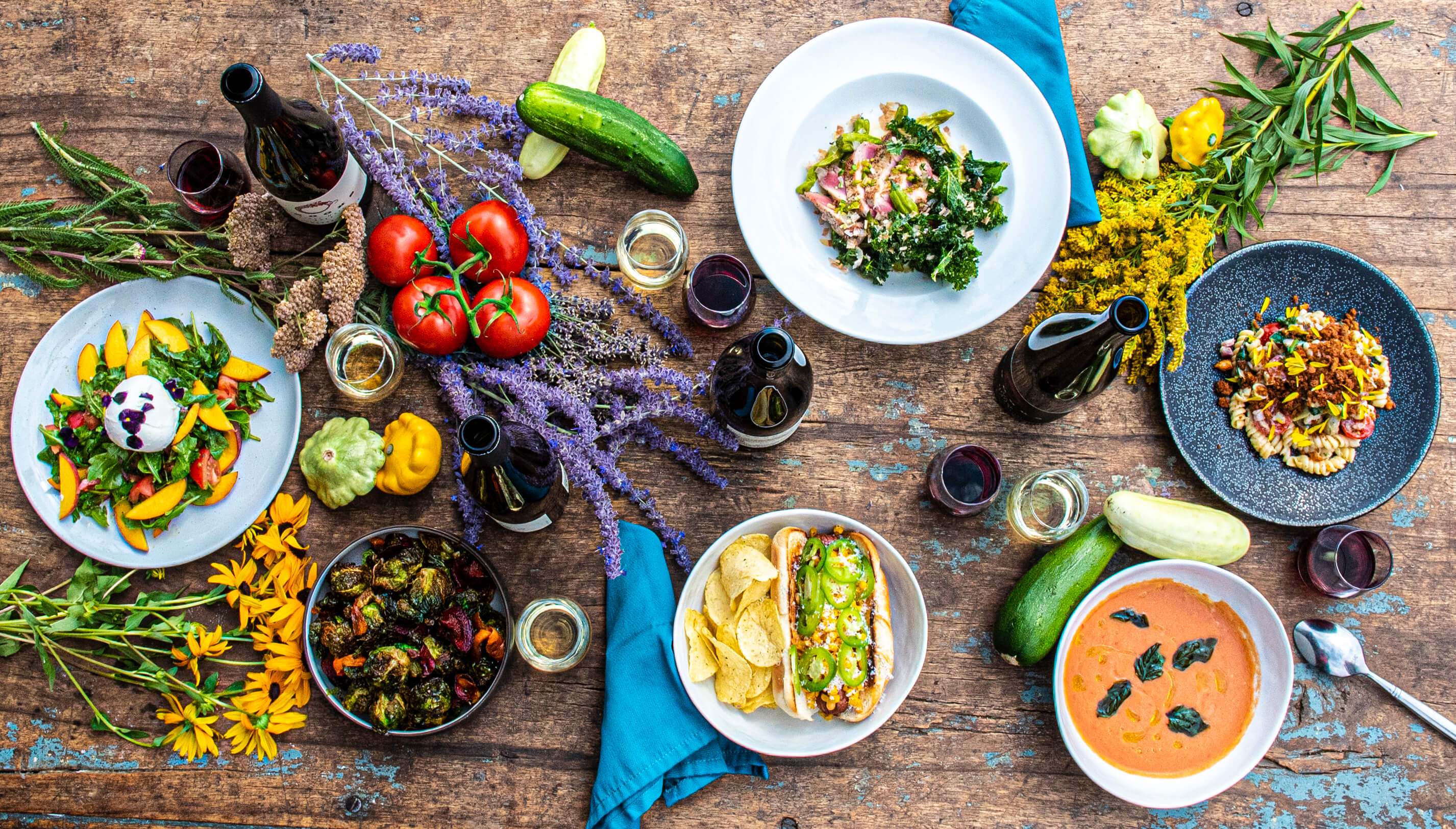 A rustic wooden table is set with flowers decorating a wine and food pairing lunch including a peach and burrata salad, tomato bisque, elote hot dog, fried brussels sprouts, and a macaroni salad with crispy shallots.