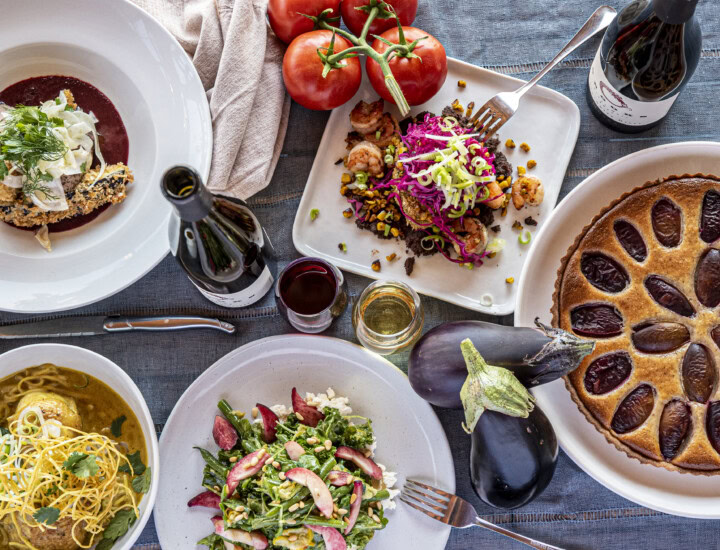 Light blue table cloth set with a wine and food pairing lunch including a plum tart, string bean salad with feta cheese, corn and corn fritter, and crispy pork loin with shaved fennel.
